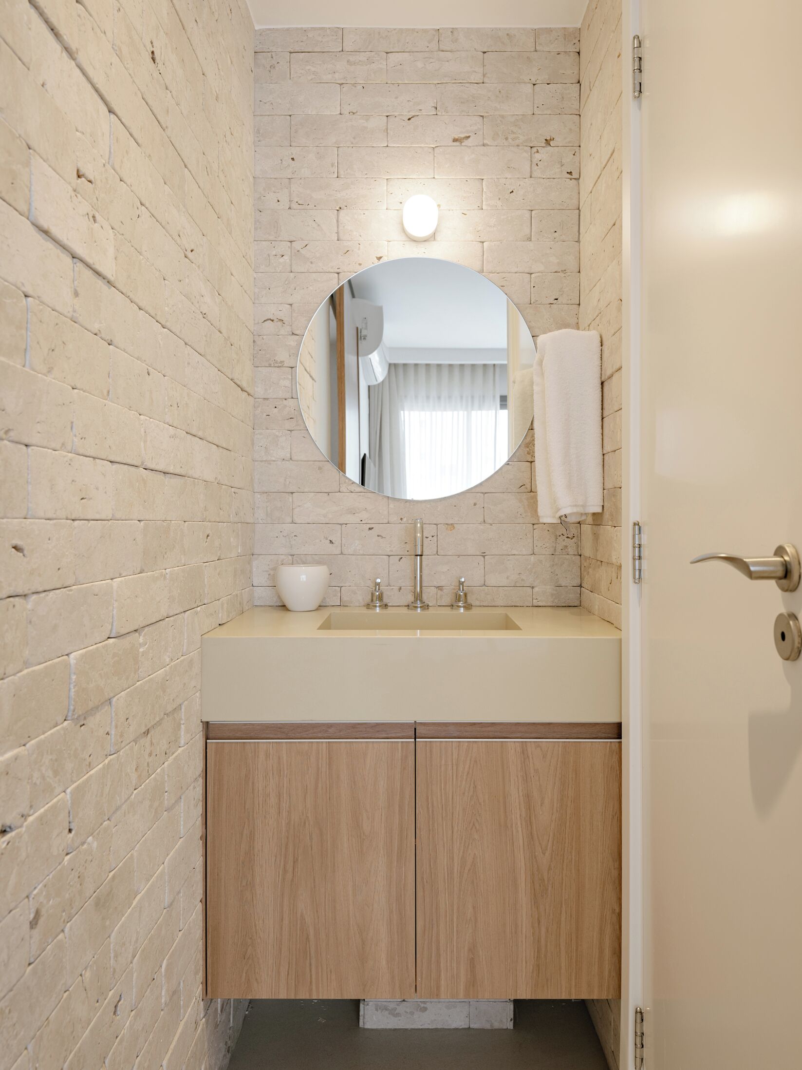 Une salle de bain avec un mur en briques de couleur beige, un miroir rond, un lavabo avec un plan de travail blanc et des meubles en bois.