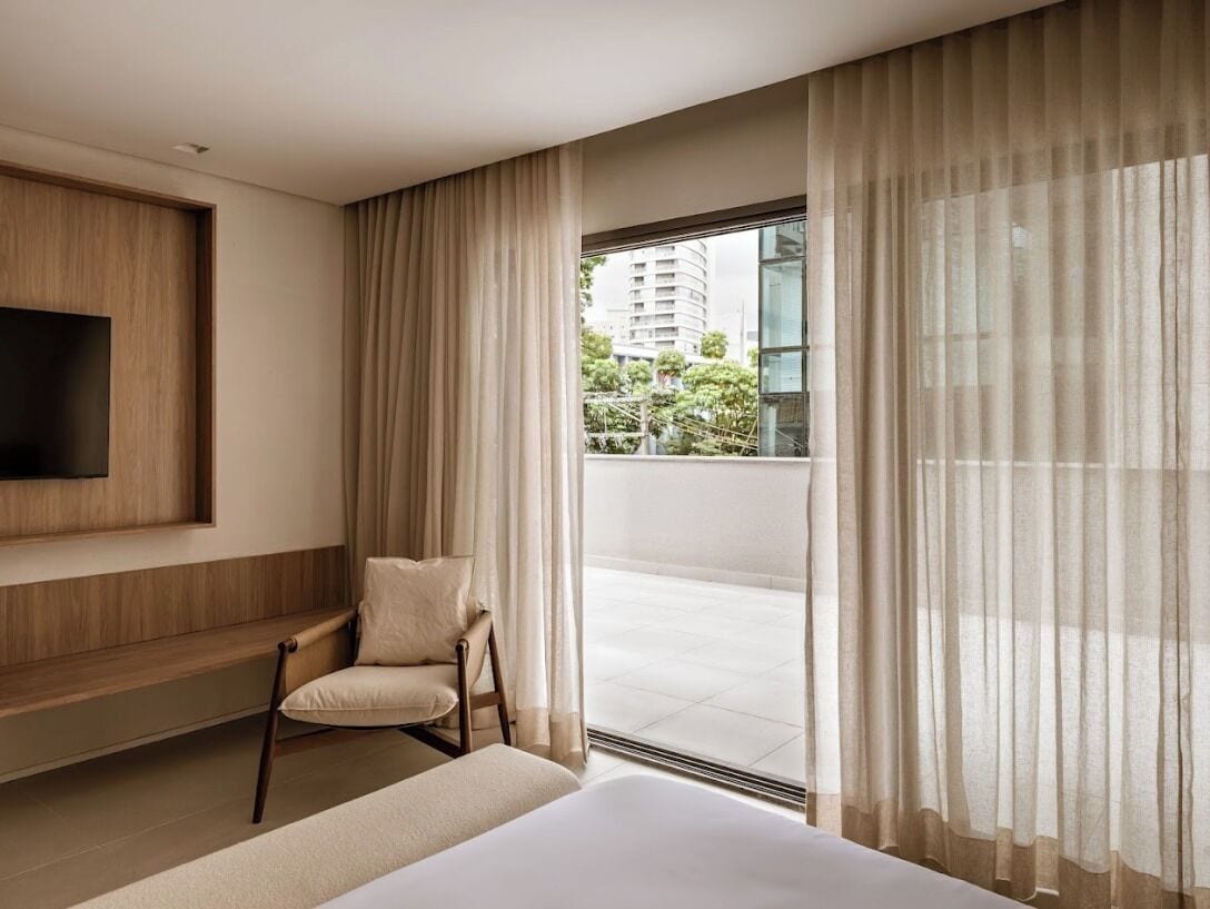 Habitació d'hotel moderna amb un llit gran, zona de seients amb una cadira, televisió de pantalla plana penjada a la paret i un balcó amb vista a edificis i verdura.