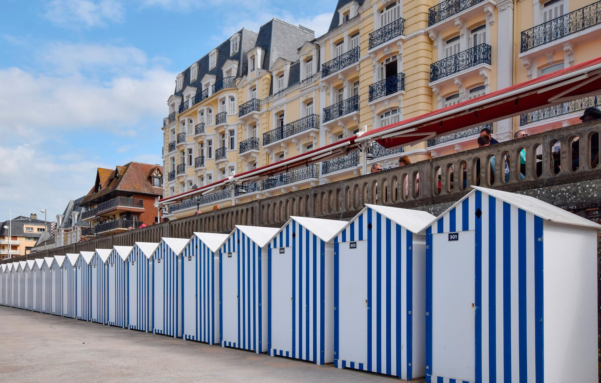 A row of blue and white striped beach huts numbered 301 to 305, with a historic building featuring balconies in the background.