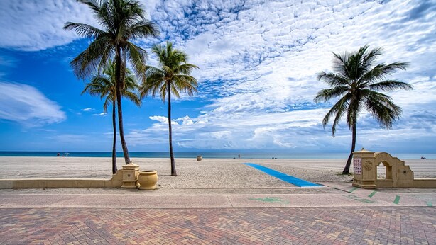 Uma praia com palmeiras, um caminho de azulejos e céu claro.
