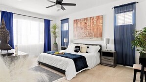 Un dormitorio moderno con cama, ventilador de techo, mesita de noche, lámpara y un ventanal con cortinas.