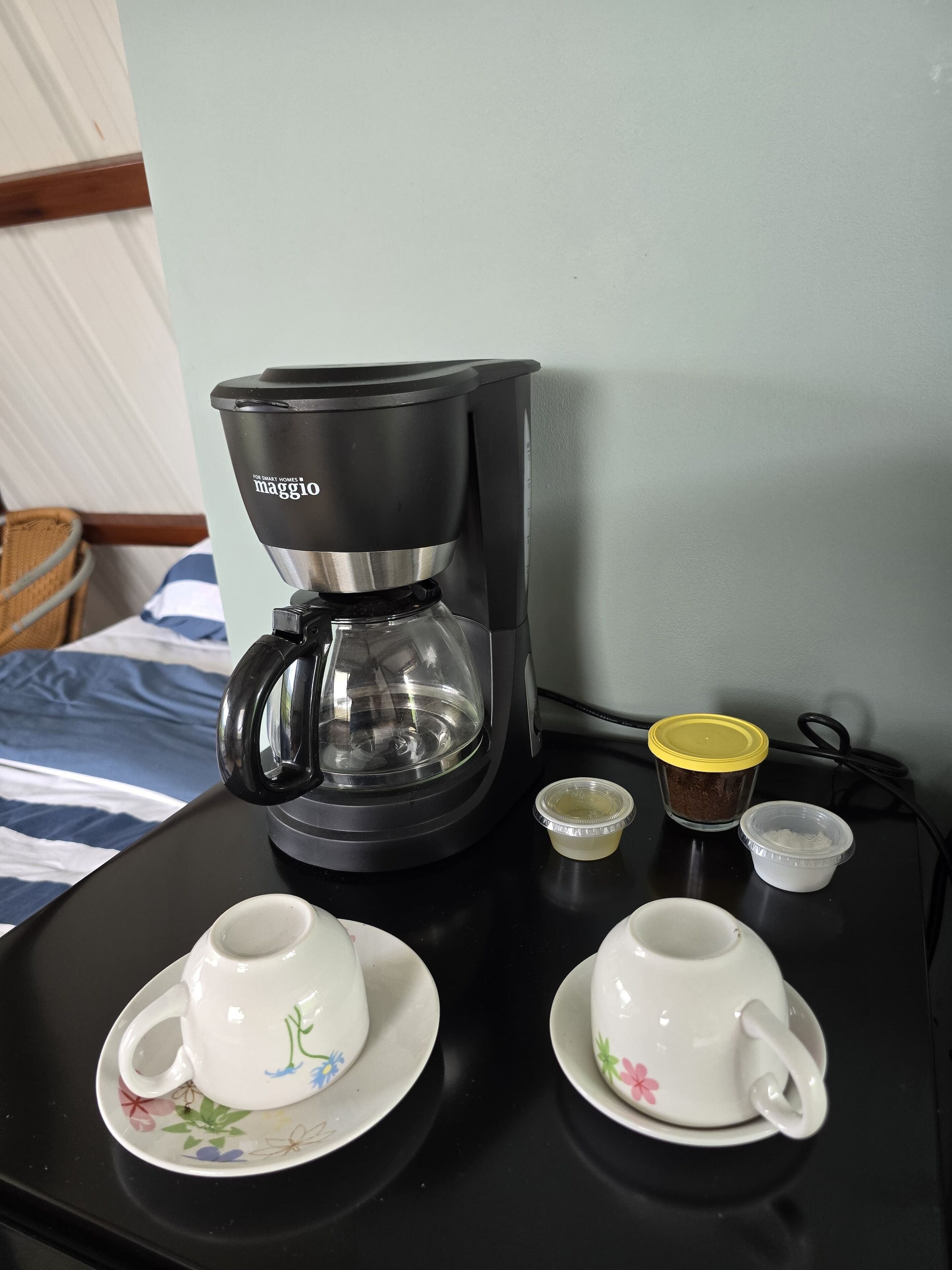 A coffee maker on a table with two cups and two small containers.