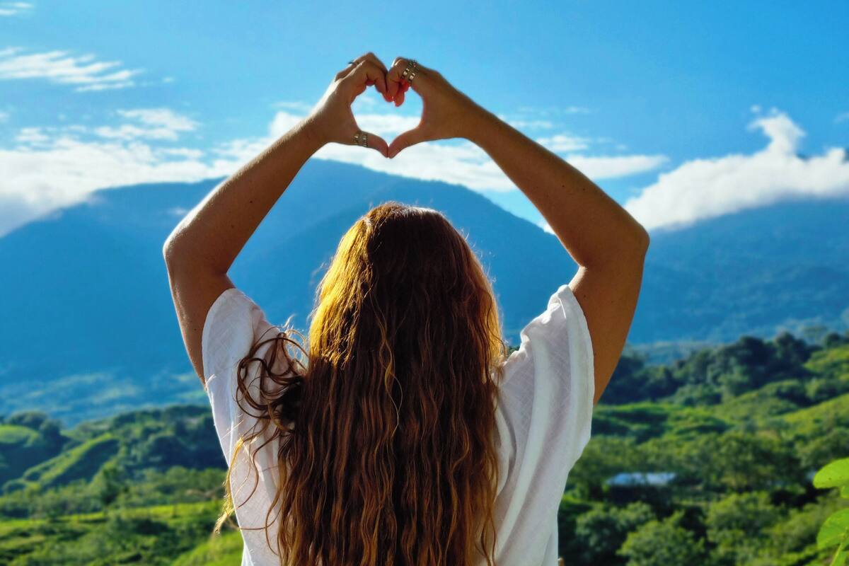 Una persona forma un corazón con las manos, de pie en un balcón con vistas a colinas verdes y montañas.