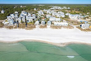 Eine Luftaufnahme einer Küstenwohngegend mit mehreren zweistöckigen Häusern, einem Sandstrand und klarem türkisblauem Wasser.