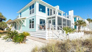 Een tweelaags strandhuis met een balkon, witte relingen en een zandstrand op de voorgrond.