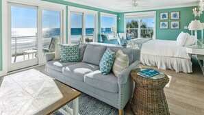 Een ruime kamer met een groot raam dat uitkijkt op het strand, een comfortabele bank met gestreepte kussens, een nachtkastje en een rieten mandje.