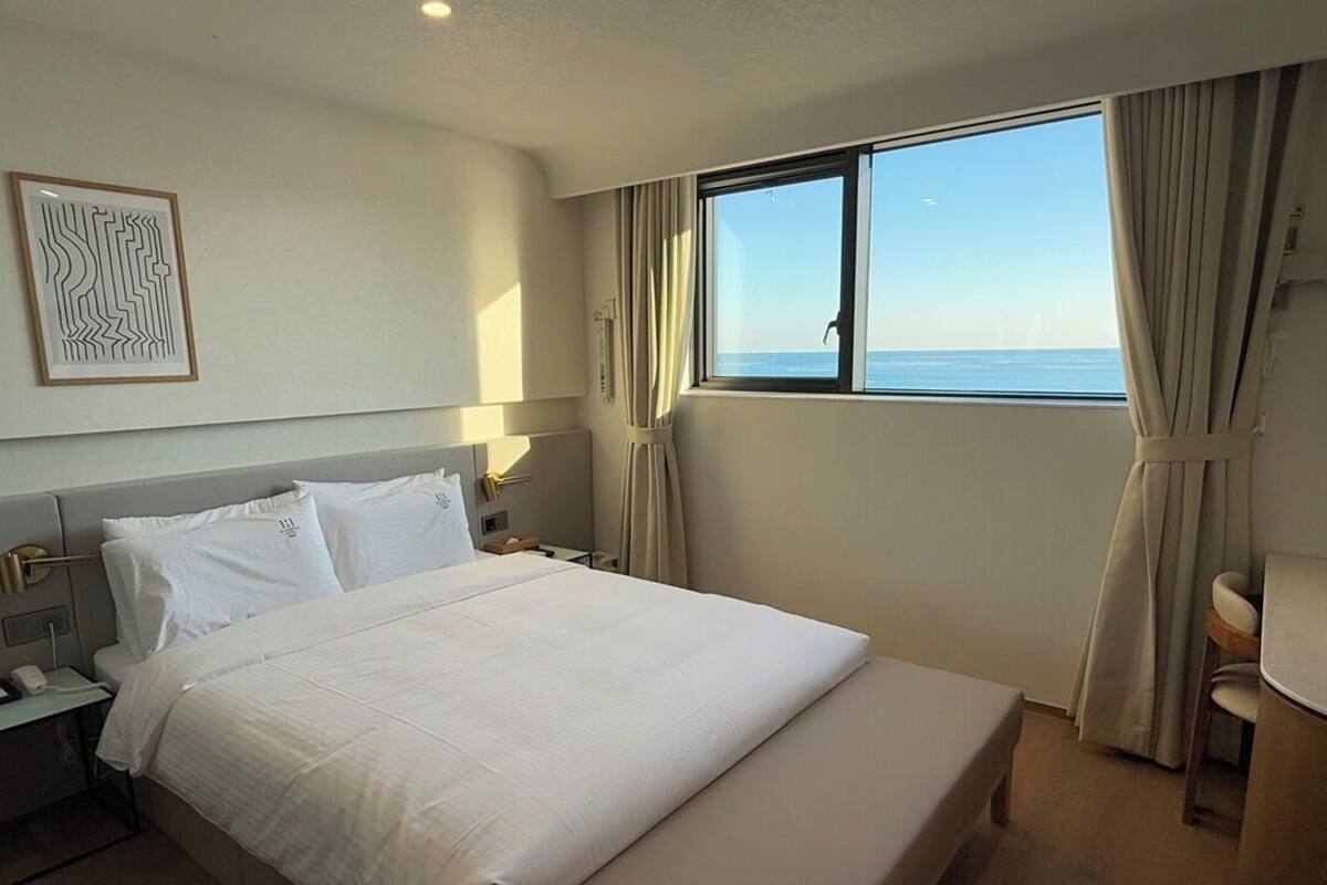 A hotel room with a large bed, a bedside table, a nightstand, a window with curtains, and a view of the sea.