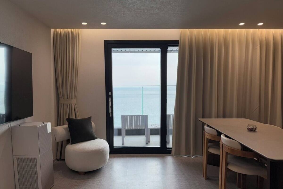 A modern hotel room with a balcony, a flat-screen TV, a compact sofa, a dining table, and a built-in air conditioning unit.