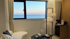A modern hotel room with a curved sofa, a small round table with amenities, a desk with a lamp, and a large window offering a view of the sea.
