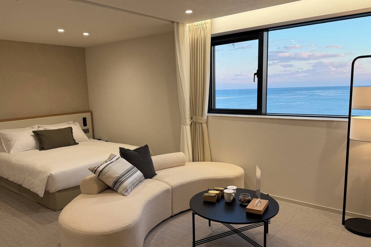 A hotel room with a bed, a sofa, a small table with items, and a window with a view of the sea.