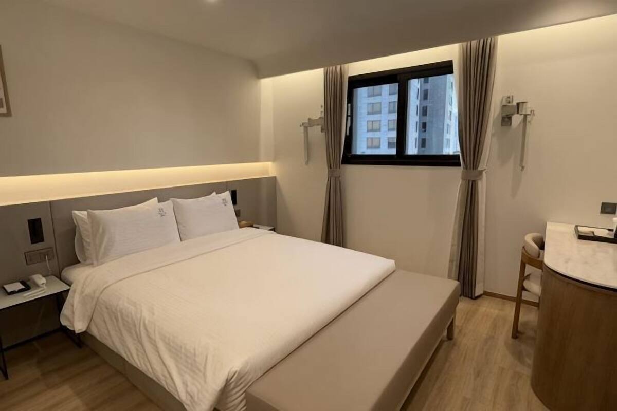 A hotel room with a bed, bedside tables, a window with curtains, a small desk with a chair, and a bathroom area.