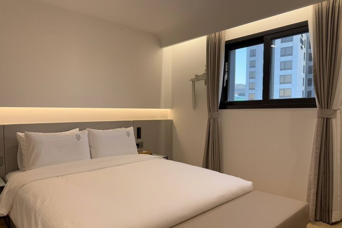 A hotel room with a large bed, a nightstand, a window with curtains, and a view of buildings outside.