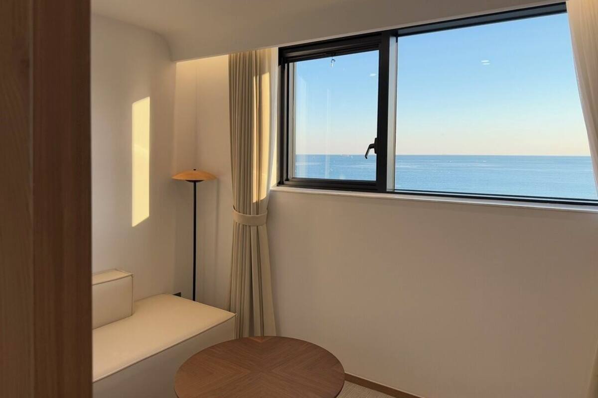 A room with a view of the sea, a round wooden table, a sofa, and a floor lamp.