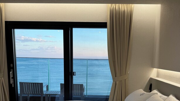 A hotel room with a bed, white pillows, and a view of the ocean through a sliding glass door.