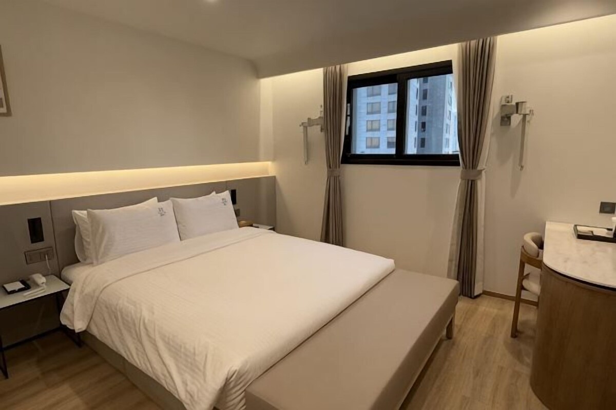A hotel room with a bed, bedside tables, a window with curtains, a small desk with a chair, and a bathroom area.