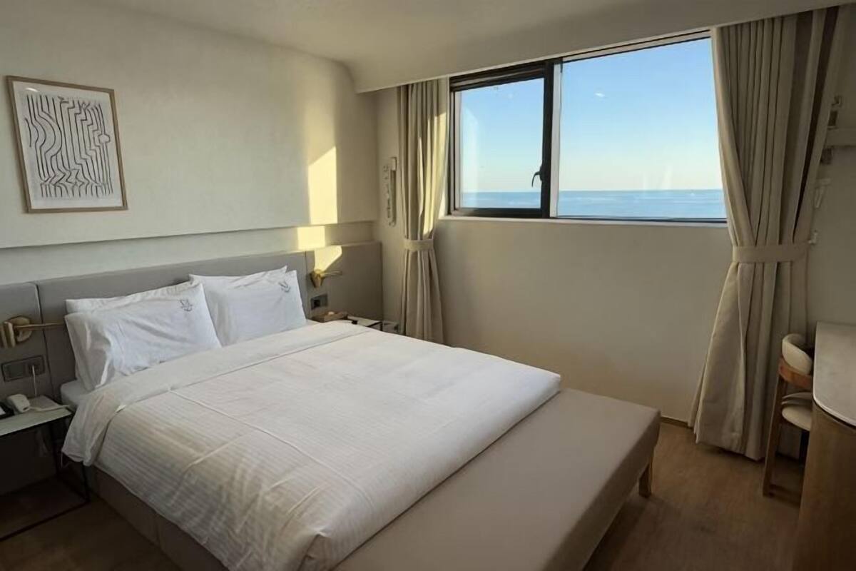 A hotel room with a large bed, a bedside table, a nightstand, a window with curtains, and a view of the sea.