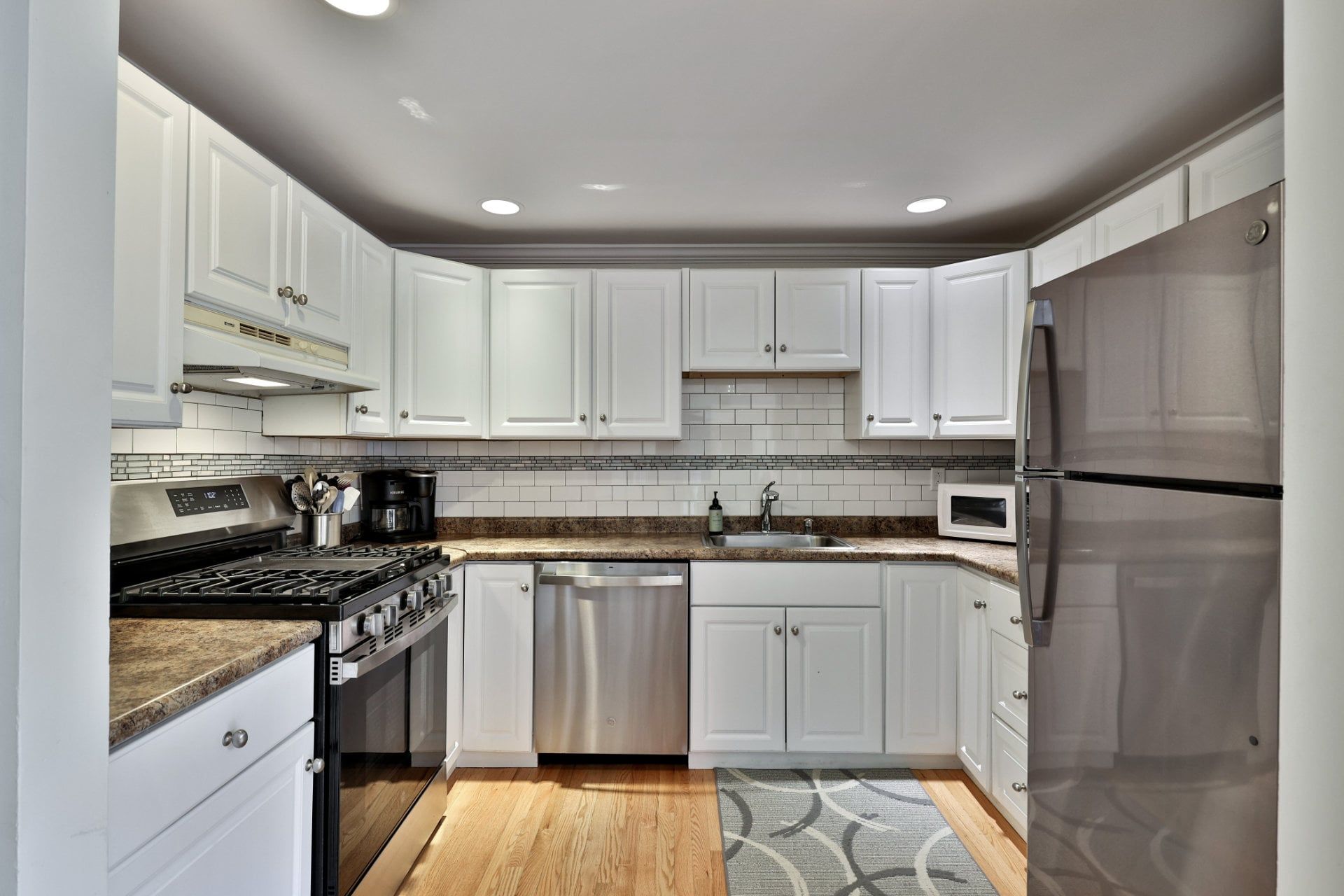 A modern kitchen with white cabinets, stainless steel appliances, and a patterned rug.