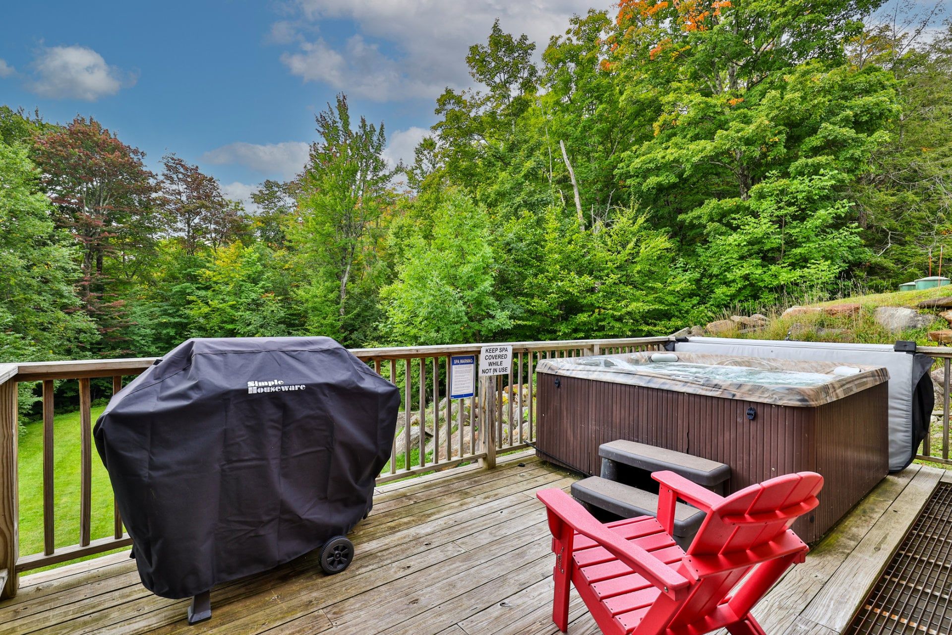 A covered grill, a hot tub, and a red chair on a wooden deck with a view of trees.