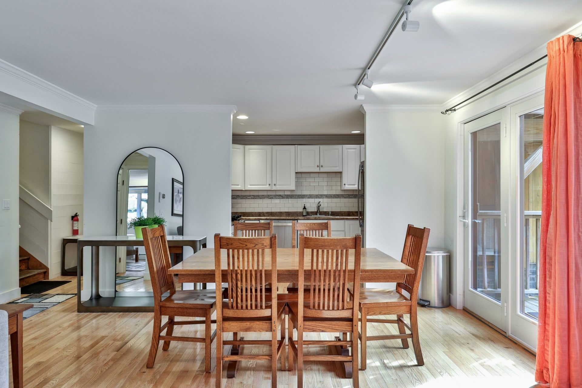 A dining area with a wooden table and chairs, a kitchen in the background, and a sliding glass door with curtains.