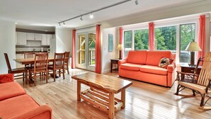A spacious living area with a wooden dining table, a sofa, and a rocking chair. - Trailside Village #1: Slopeside 1BR w/ Hot Tub and Sauna. Ski In/Out with Discounted Ski Rentals (Killington)