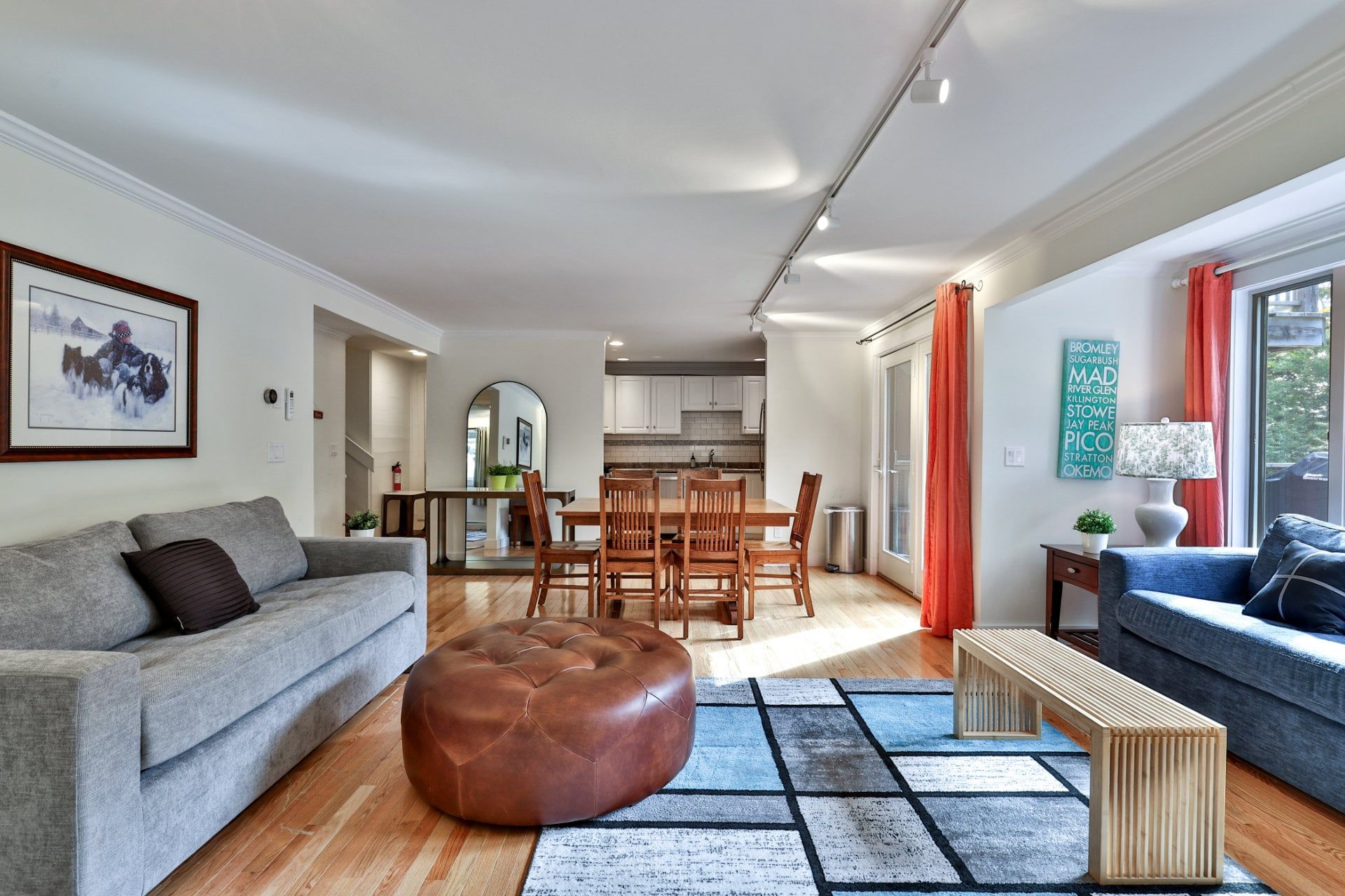 Una sala de estar moderna con un sofá gris, un reposapiés marrón y un sofá azul. Hay un comedor con sillas de madera y una mesa, y una cocina al fondo.