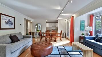 Una sala de estar moderna con un sofá gris, un reposapiés marrón y un sofá azul. Hay un comedor con sillas de madera y una mesa, y una cocina al fondo.