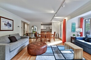 En moderne stue med en grå sofa, en brun skammel og en blå sofa. Der er et spiseområde med træstole og et bord, og et køkken i baggrunden.