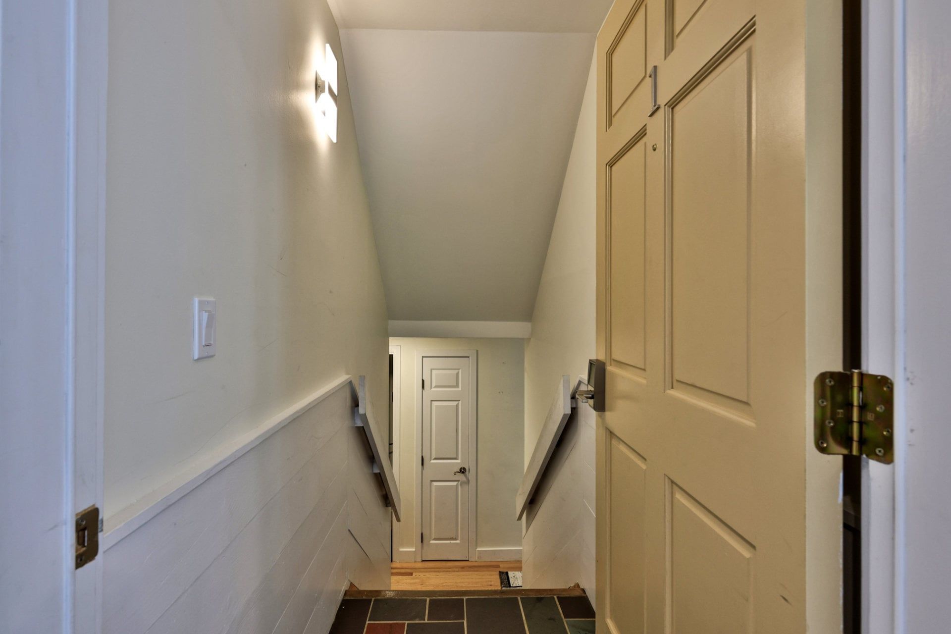 A narrow hallway with white walls, a door at the end, and a light fixture on the left.