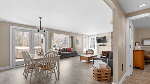 A spacious living room with a dining area, large windows, and a flat-screen TV. - Trailside Village 5: Wood Stove, Private Sauna and Hot Tub (Killington)
