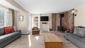 A living room with a fireplace, two sofas, a coffee table, and a TV. - Trailside Village 5: Wood Stove, Private Sauna and Hot Tub (Killington)