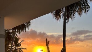 A balcony with a view of the ocean at sunset, palm trees, and a white fence. - Surfside Beach Condo in wonderful Aguada, PR (Aguada)