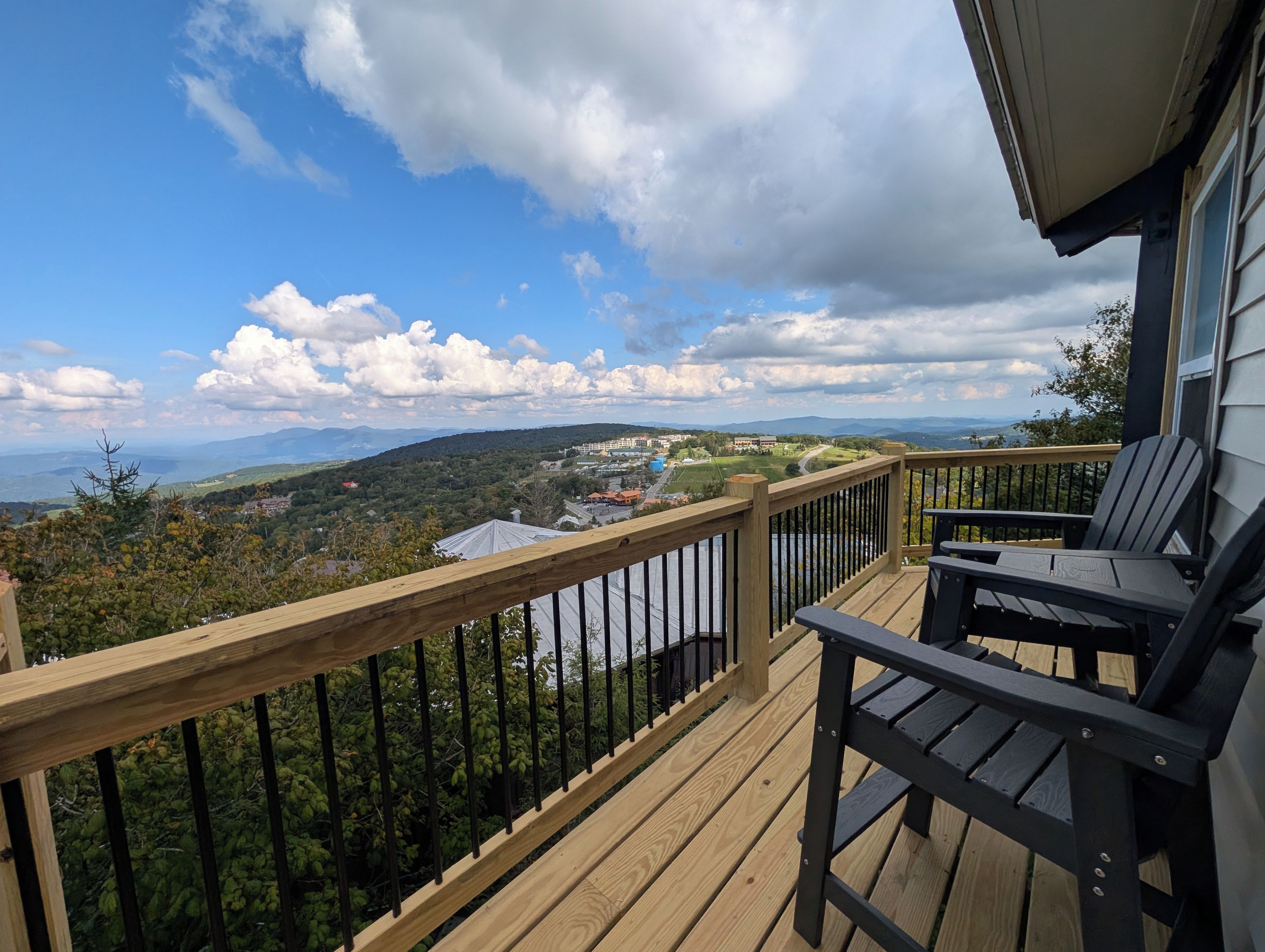Una terraza de madera con barandilla, dos sillas y vistas a un paisaje con montañas y una masa de agua.