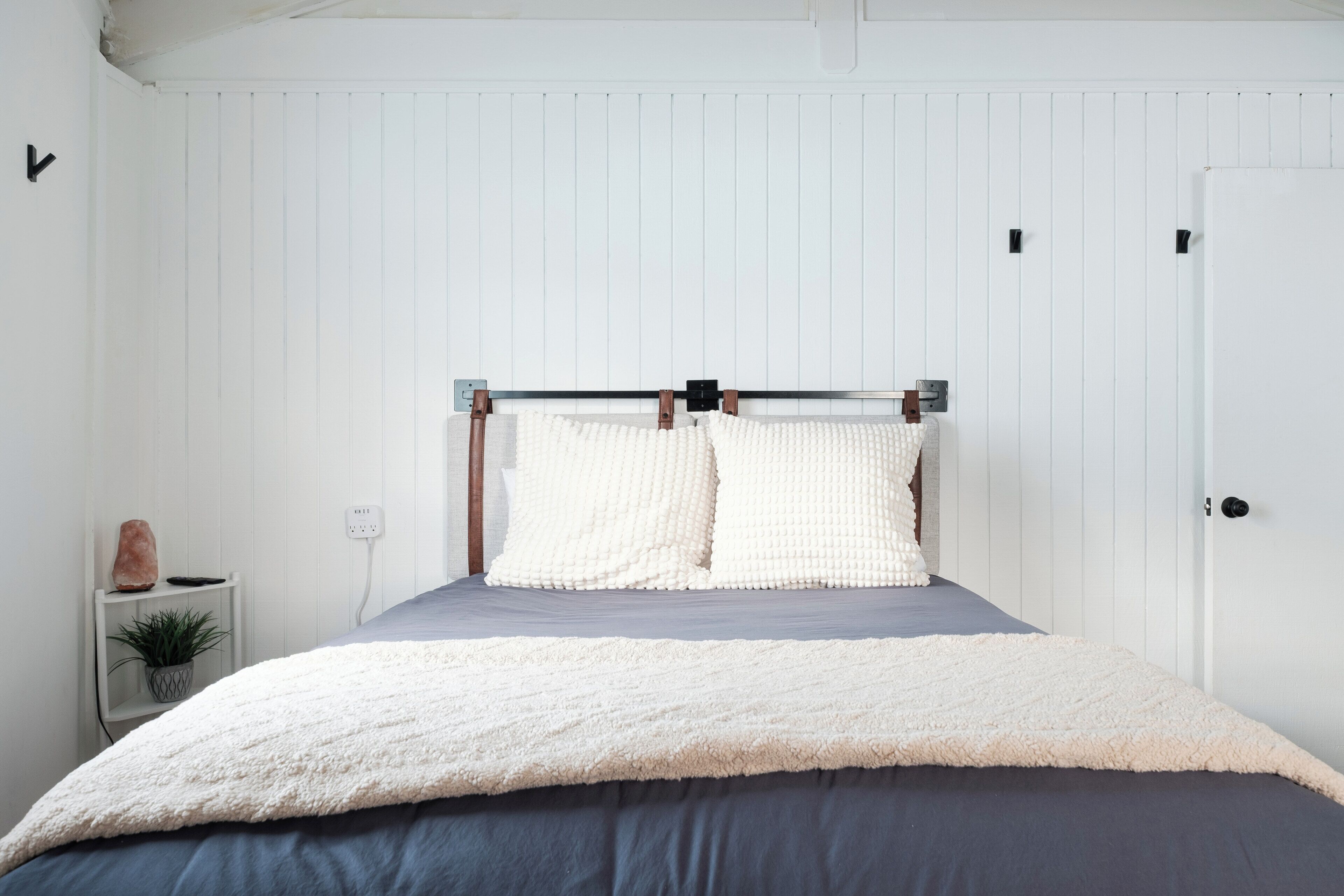 Une chambre à coucher avec un lit, des murs blancs, une petite table de chevet avec une plante et une porte.