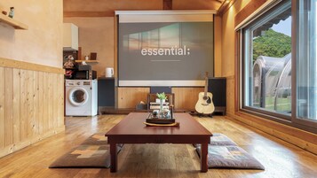 A room with a wooden floor, a projector screen displaying 'essential;', a washing machine, a guitar, and a table with a tray.