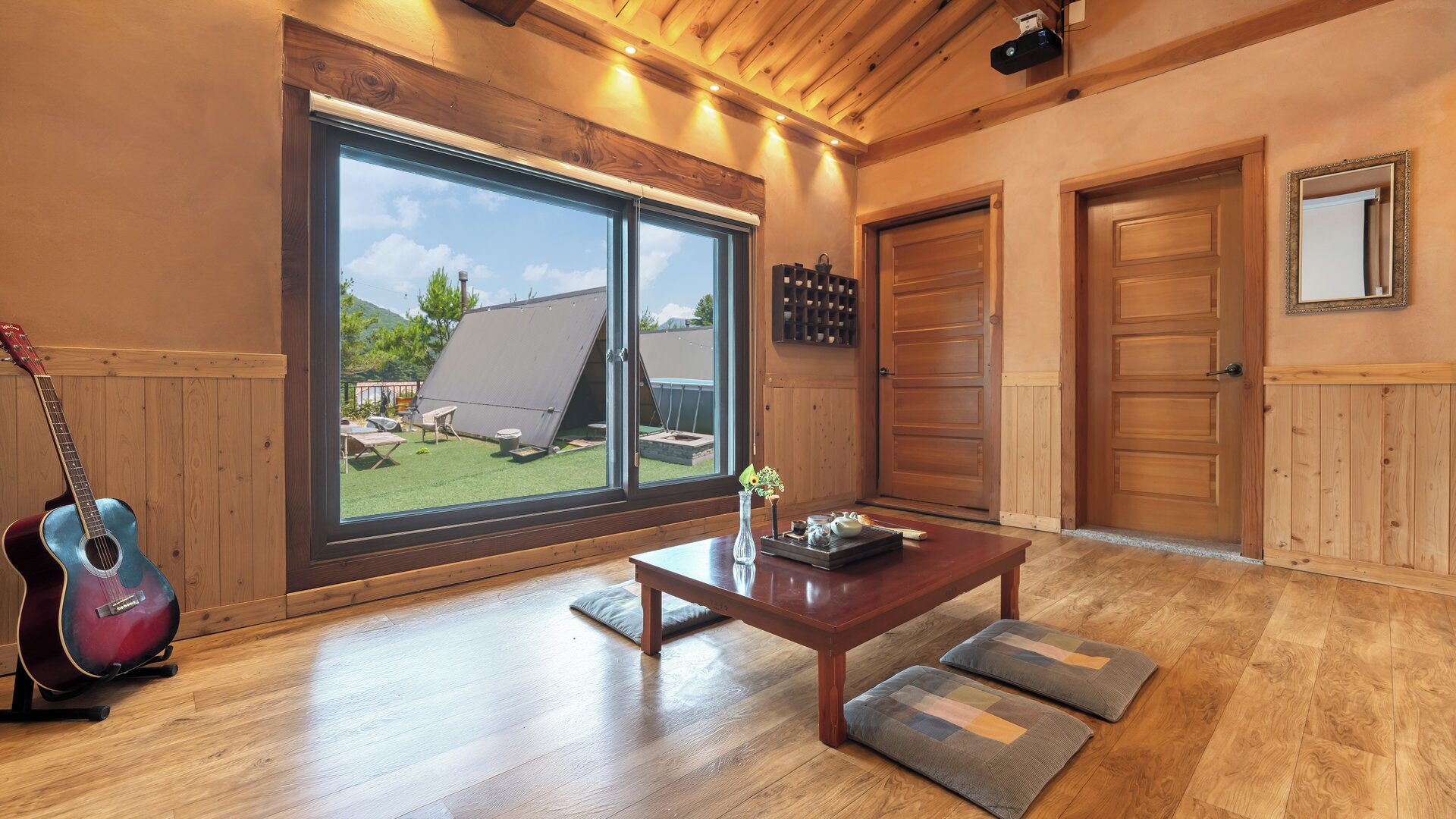 A room with a large sliding glass door, a wooden ceiling, a guitar, a table with cushions, and a view of a house and trees.