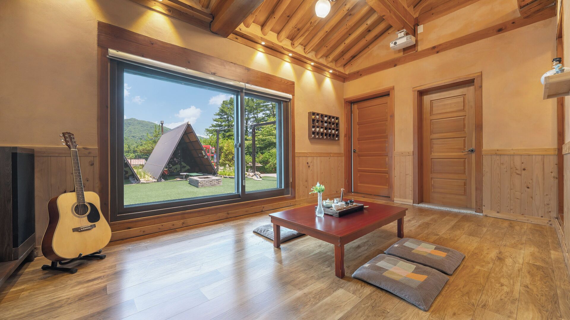 A room with a wooden floor, a guitar, a table with a vase, and a view of a tent outside.