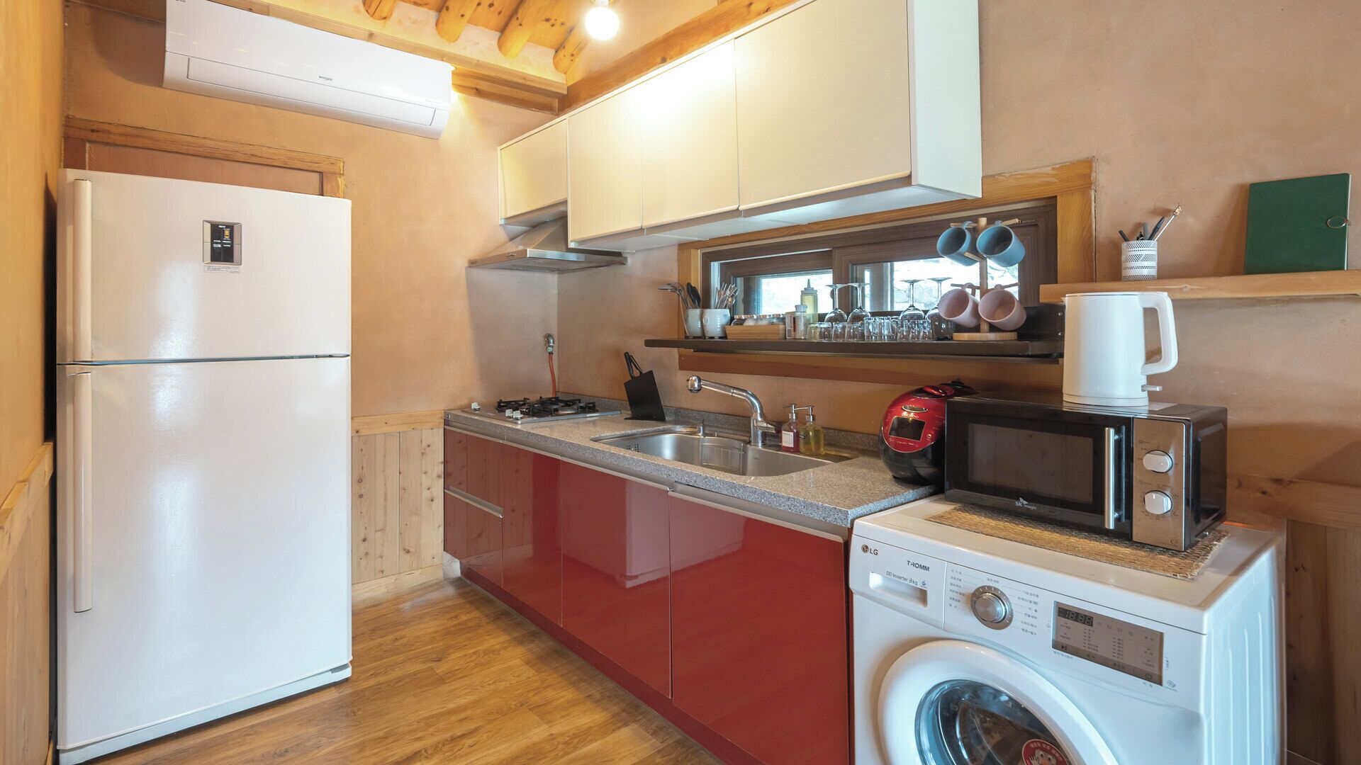 A compact kitchen with a refrigerator, microwave, and washing machine, wooden cabinets, and a window above the sink.