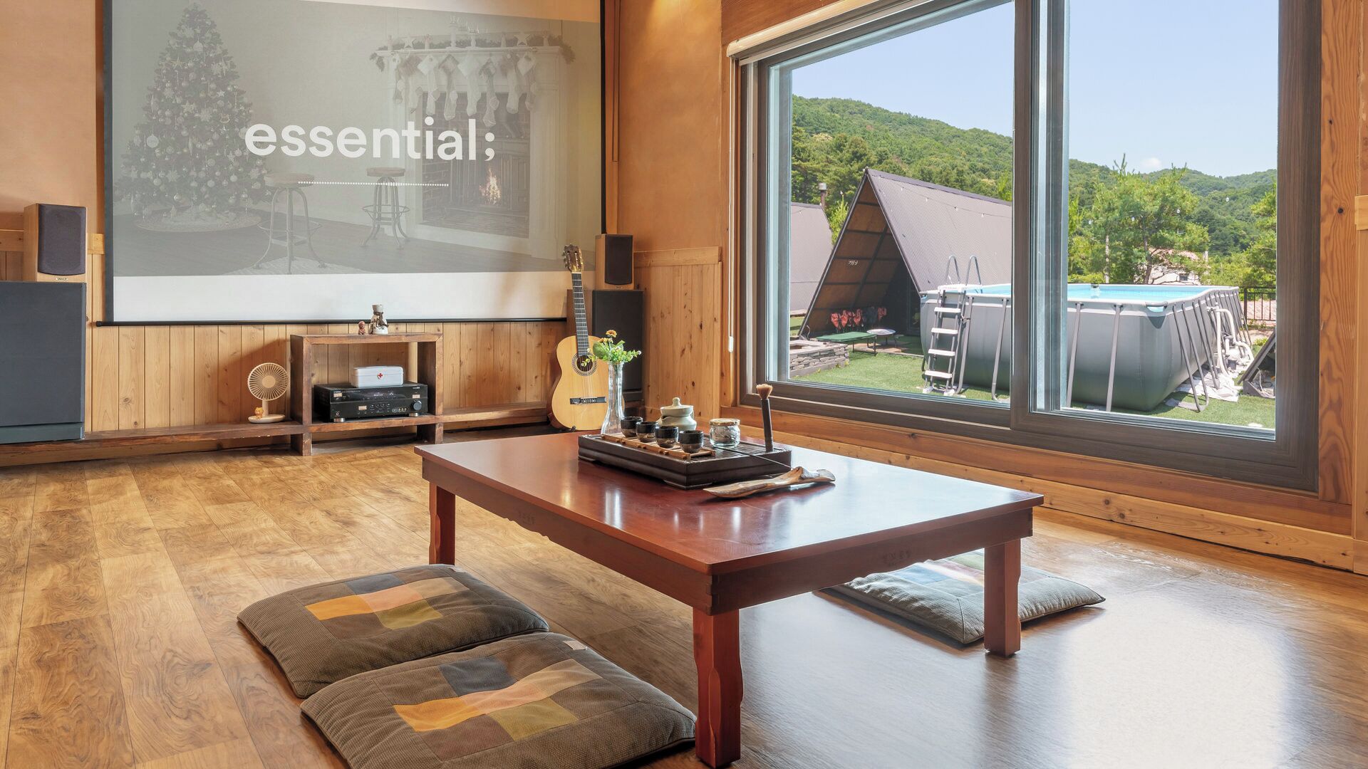 Una habitación acogedora con un ventanal grande, una mesa de madera y una pantalla de proyector que muestra la palabra 'essential'.