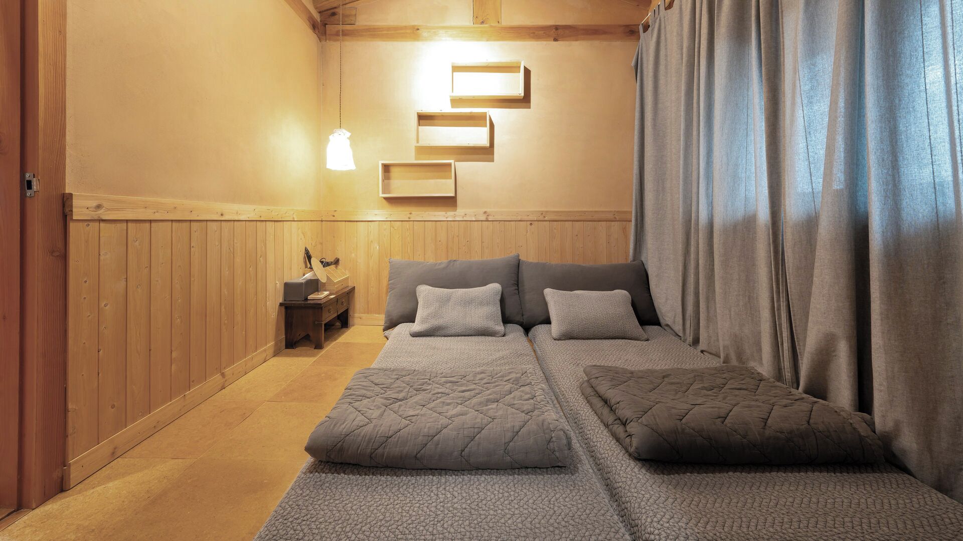 Una habitación con piso de madera, una cama con cubrecama acolchado, una mesita de noche con lámpara y un estante fijado a la pared.