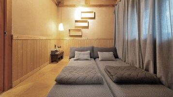 A room with a wooden floor, a bed with a quilted bedspread, a bedside table with a lamp, and a wall-mounted shelf.