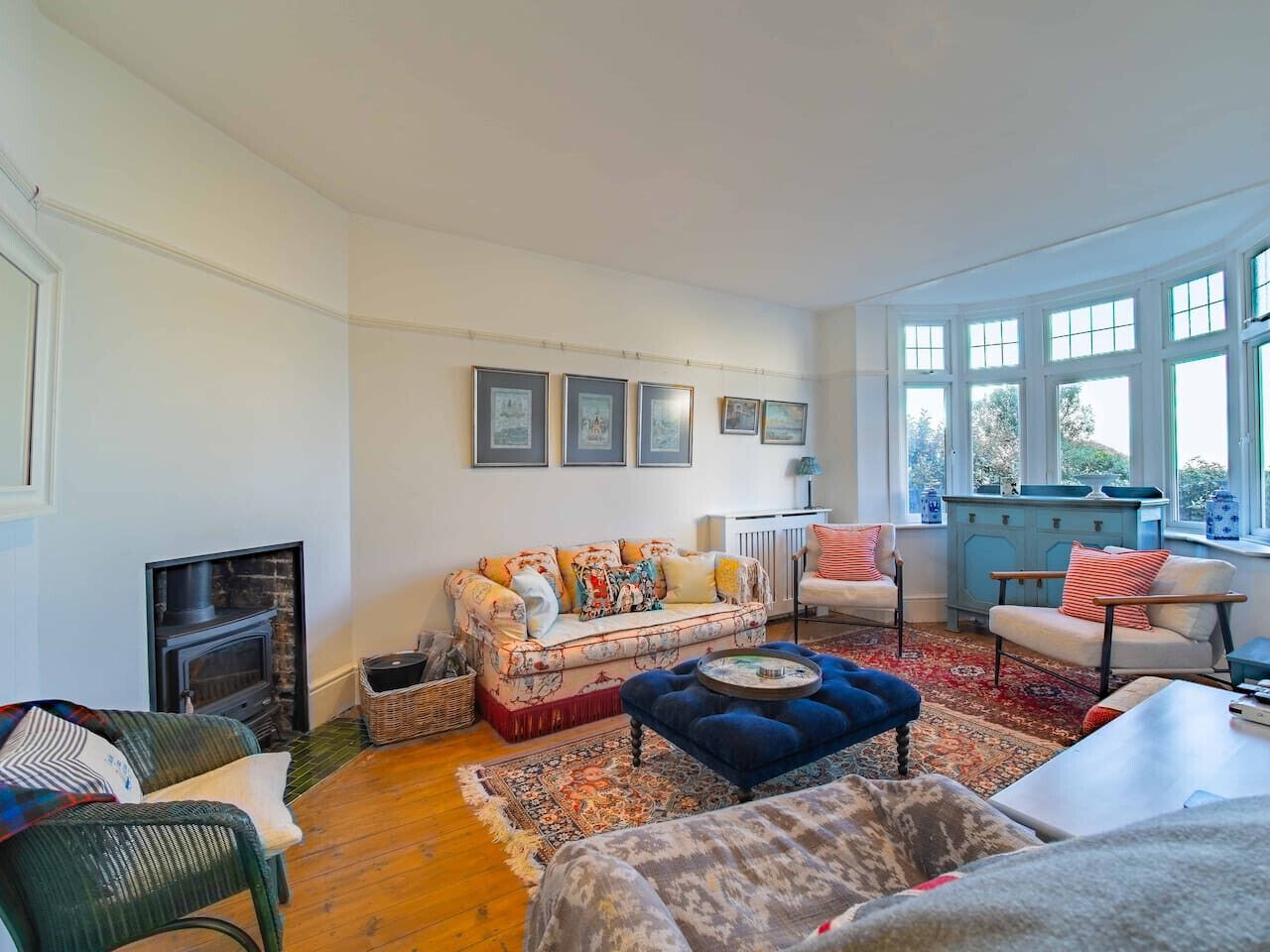 A cozy living room with a fireplace, a sofa, a blue ottoman, and a view of the outdoors through large windows.