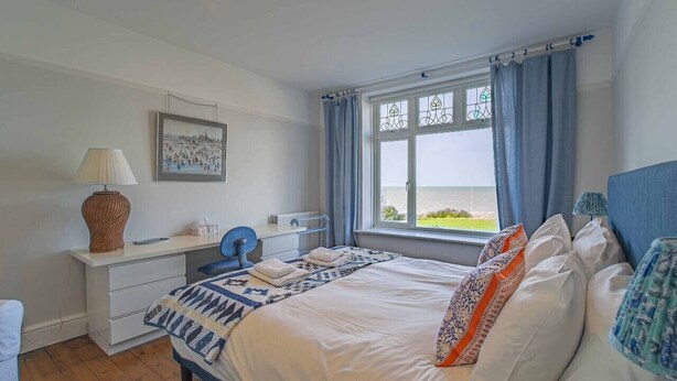 A bedroom with a bed, a bedside table, a lamp, a window with curtains, and a view of the sea.