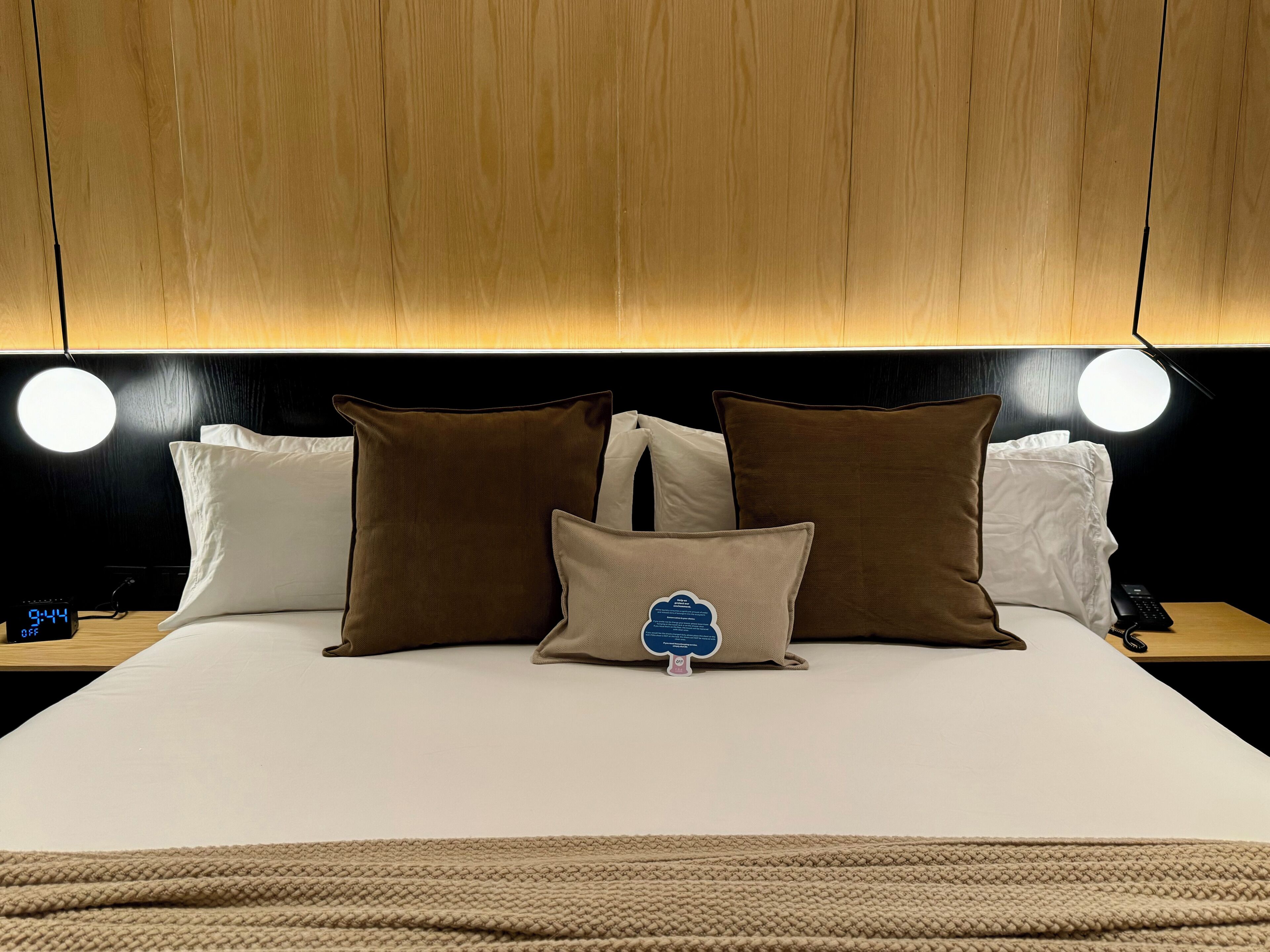 A neatly made bed with a white mattress, brown and white pillows, and a knitted throw blanket. There are two bedside tables with lamps and a digital clock displaying the time.