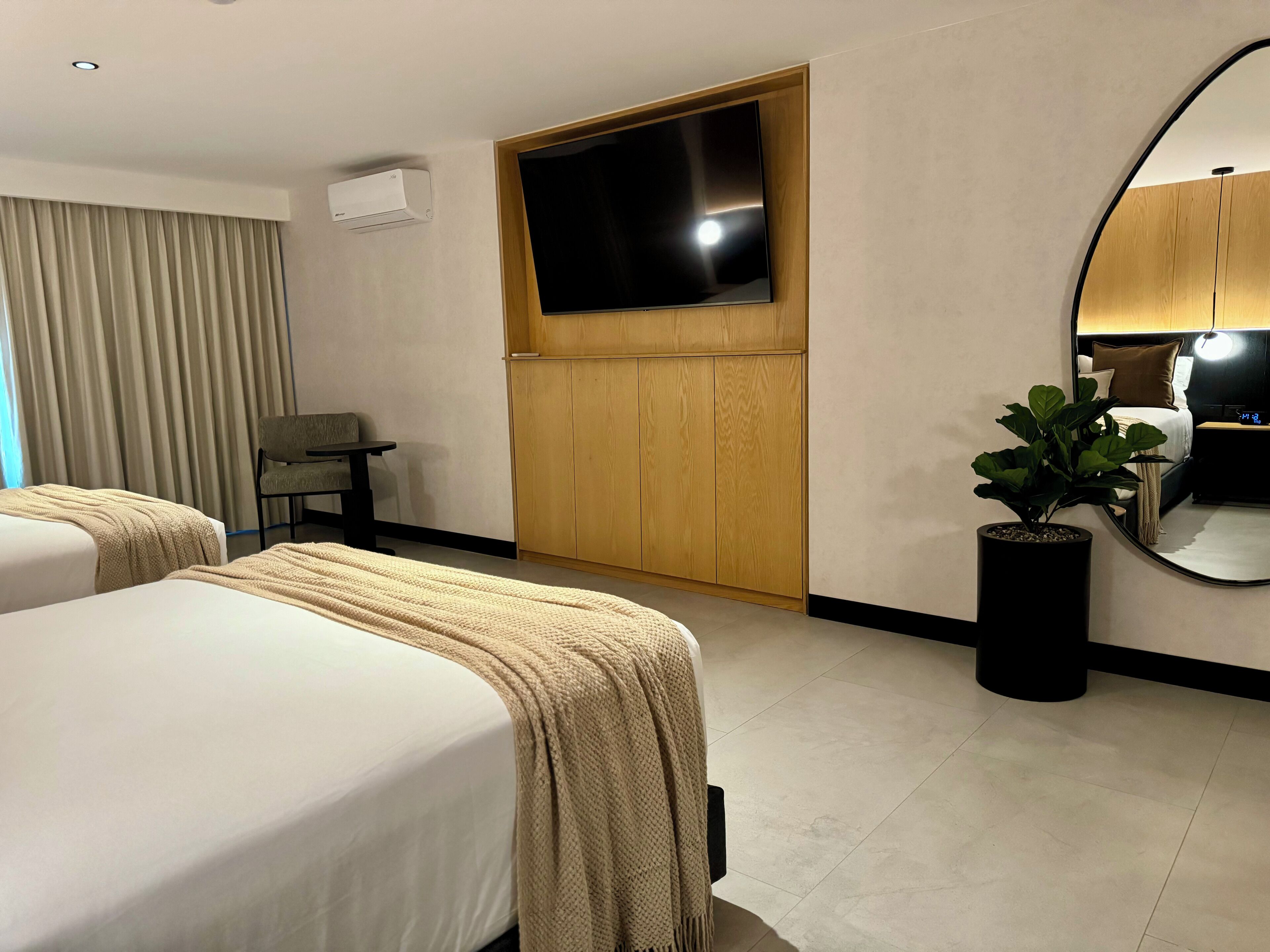 A hotel room with two beds, a TV mounted on the wall, a wooden wardrobe, a small table, and a potted plant.