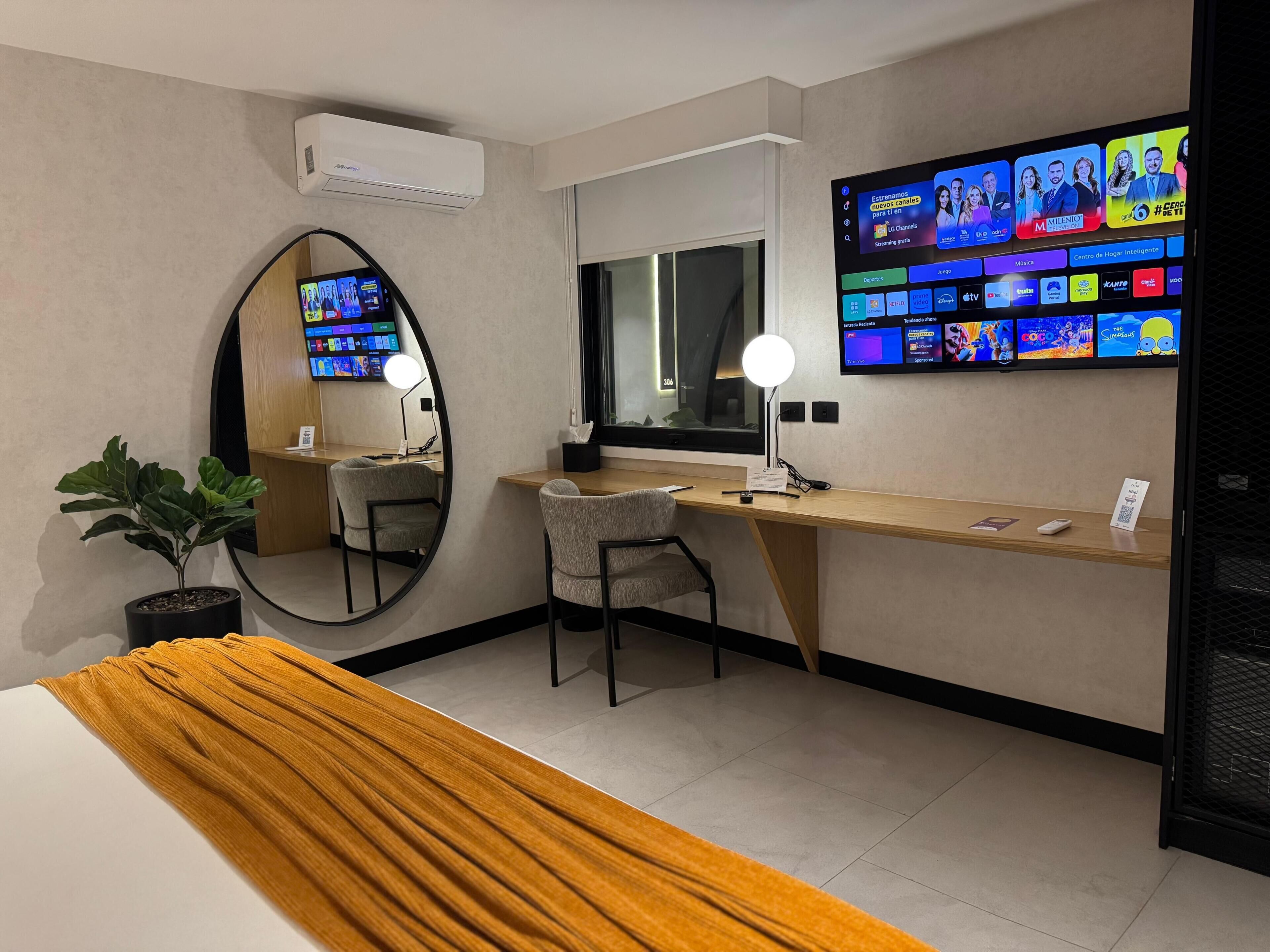 Ett hotellrum med en säng, ett skrivbord, en stol, en stor spegel, en tv som är monterad på väggen och en krukväxt.