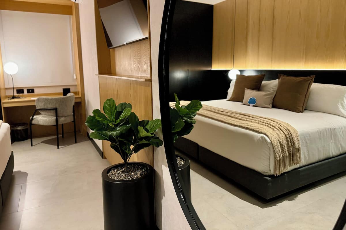 Une chambre d’hôtel moderne, équipée d’un lit, d’une table de chevet et d’un bureau avec une chaise. Une plante en pot est posée sur le sol.