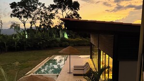 Un área de piscina moderna al aire libre con sillones de descanso y una sombrilla de paja, rodeada de vegetación exuberante y vistas a montañas distantes.