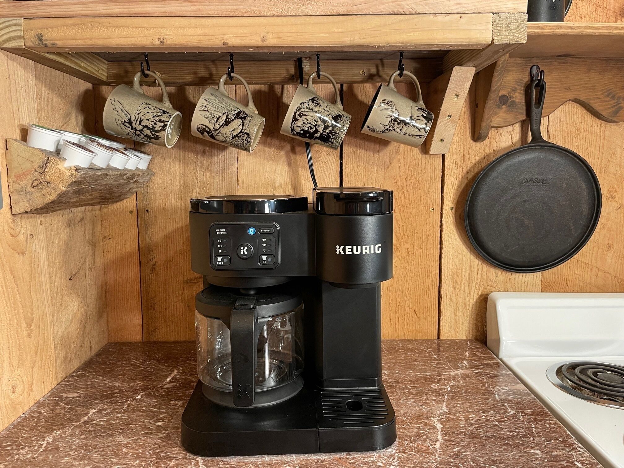 Una cafetera Keurig sobre una encimera de cocina, con un estante de madera encima que contiene tazas.