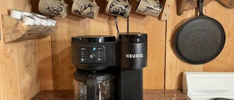 Una cafetera Keurig sobre una encimera de cocina, con un estante de madera encima que contiene tazas.