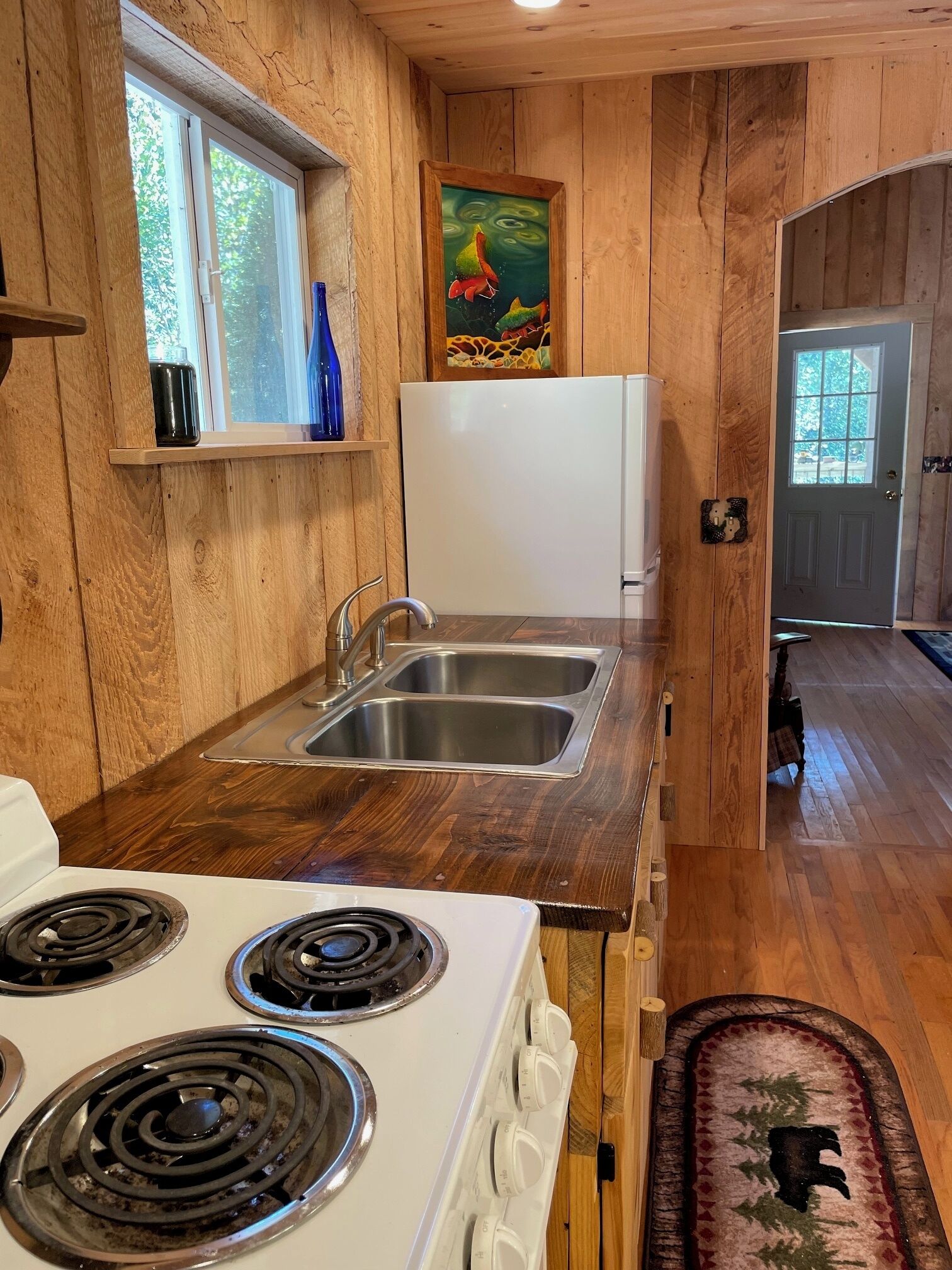 A compact kitchen with wooden walls, a white stove, stainless steel sink, and a window with a view.